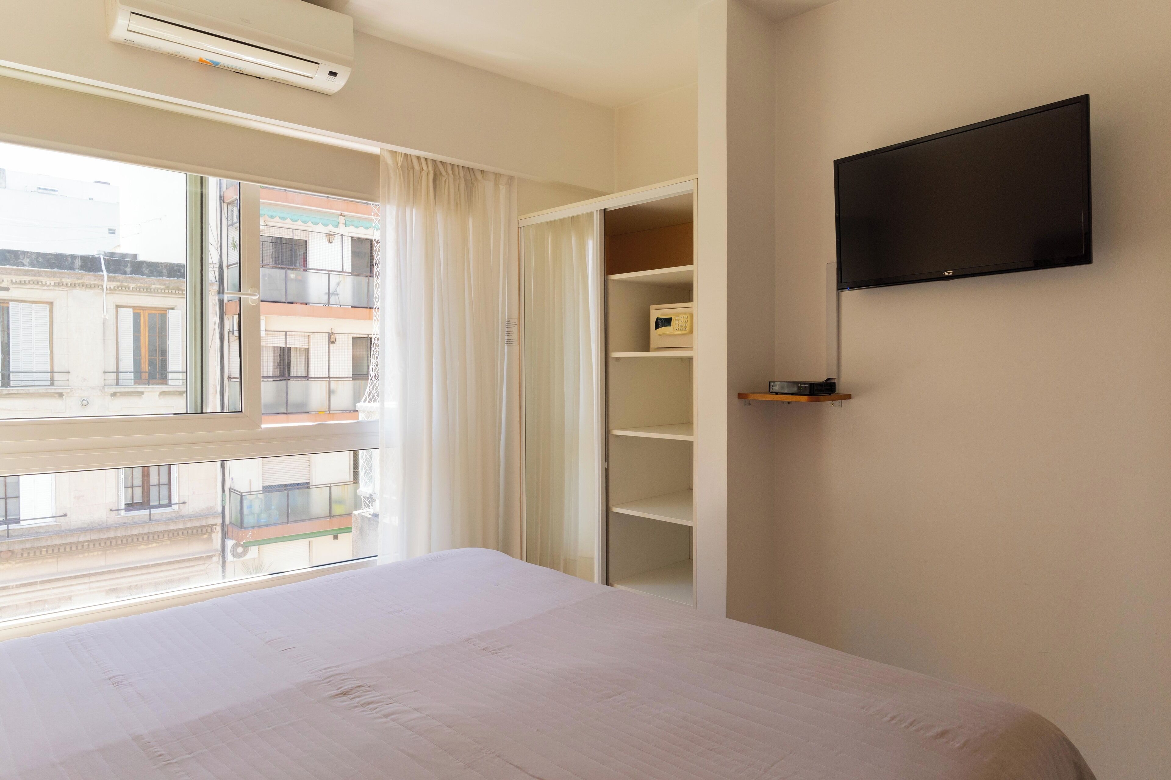 Een hotelkamer met een bed, een aan de muur bevestigde televisie, een raam met gordijnen en een uitzicht op gebouwen.