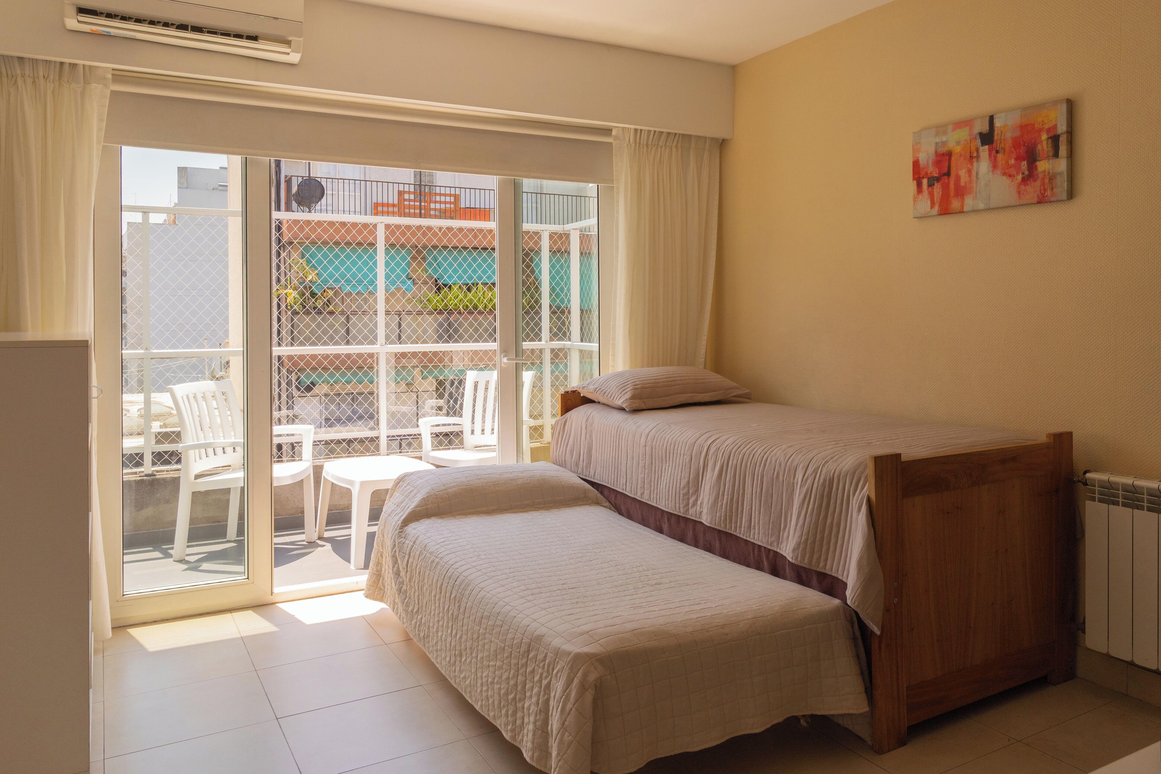 A bedroom with a bed, a bench, a sliding glass door to a balcony, and a painting on the wall.