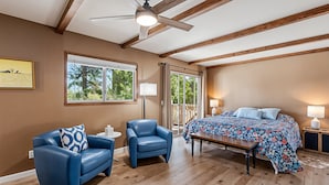 Une chambre avec un plafond en bois, un lit recouvert d’une couvre-lit à motifs floraux, un fauteuil bleu, une petite table et une fenêtre donnant sur des arbres.