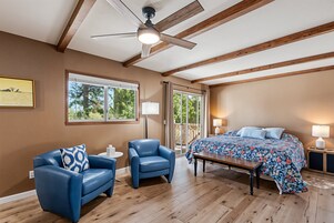 A bedroom with a wooden ceiling, a bed with a floral bedspread, a blue armchair, a small table, and a window with a view of trees.