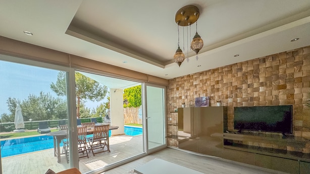 A modern living room with a large TV, a sofa, and a pool view. - Exclusive Villas (Kusadasi)