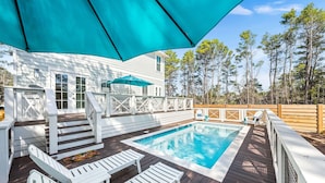 Um deck de madeira com piscina, mobiliário de exterior branco e um grande guarda-sol.