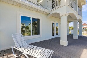 A covered outdoor area with a white lounge chair, a wooden deck, and a building with large windows and a balcony.