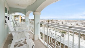 A covered balcony with white railings and lounge chairs, overlooking a sandy beach and ocean.