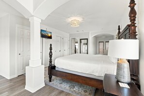 A bedroom with a large bed, a bedside table with a lamp, a television on the wall, and a view of the outdoors through a door. - ☀️Sea Forever-Walk2Beach-Stunning Home-5BR-Great Location☀️ (Pensacola Beach)