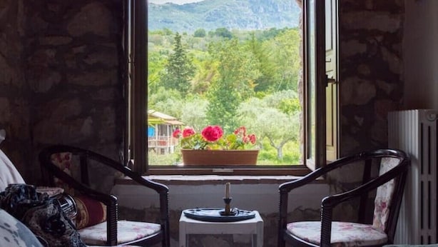 A room with a stone wall, a window with a view of greenery and mountains, two chairs, and a small table with a candle. - Villa de Luccheri - Stay in Nature (Frasso Telesino)