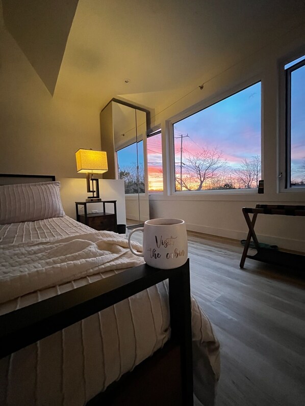 A bedroom with a bed, a bedside table with a lamp, a coffee mug, and a large window with a view of the sky.