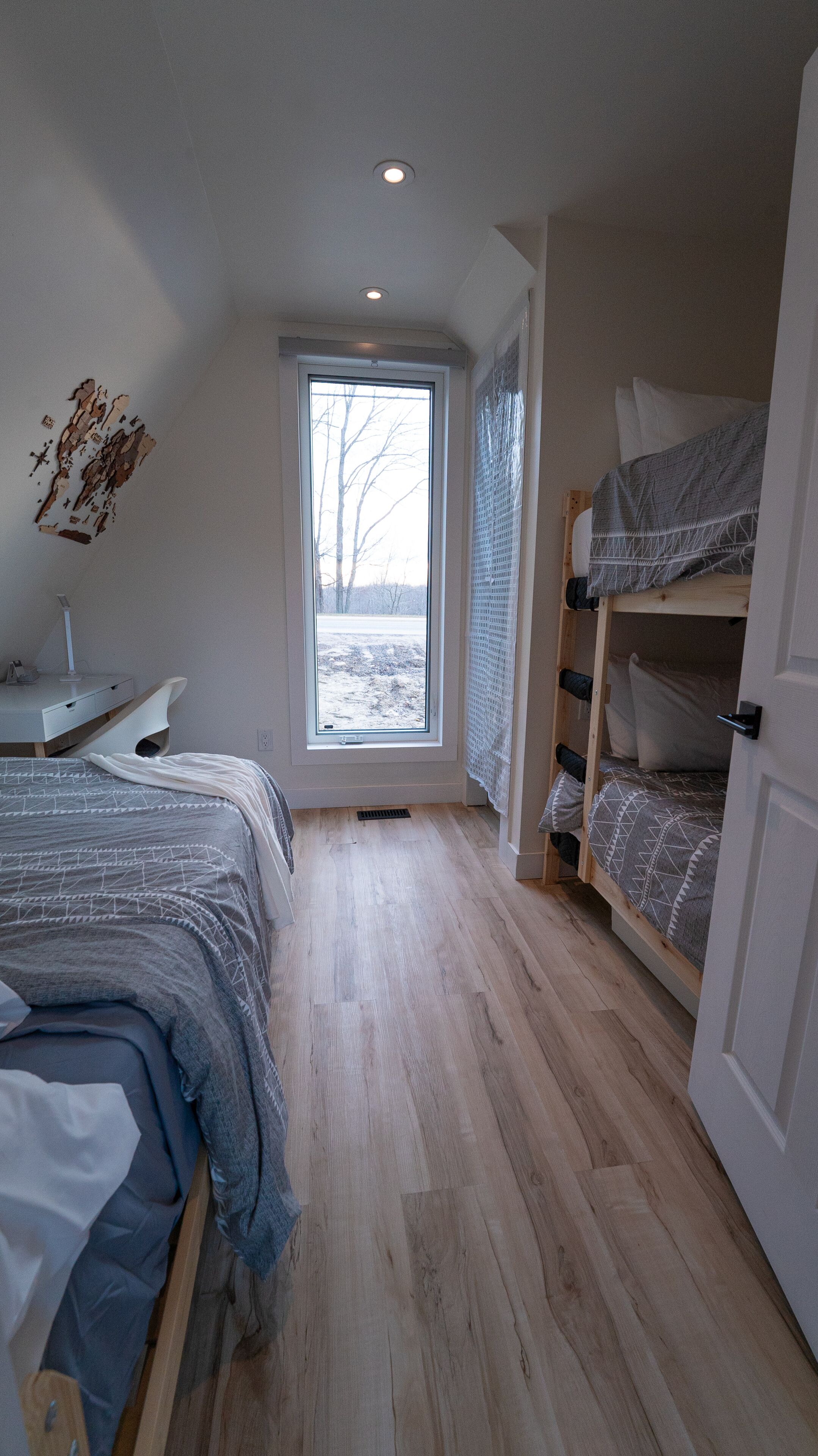 A bedroom with a bunk bed, a desk with a chair, a large window, and a ceiling light.