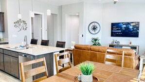 Eine moderne Küche mit einer zentralen Kochinsel, Essbereich mit Holzstühlen, einem braunen Ledersofa und einem an der Wand befestigten Fernseher.