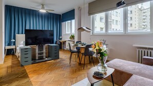 A modern living room with a flat-screen TV, a dining area with a table and chairs, a ceiling fan, and large windows with a city view. - Apartament Wiejska I (Warsaw)