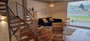Een moderne woonkamer met een trap, een bank, een fauteuil en een groot raam met zonwering.