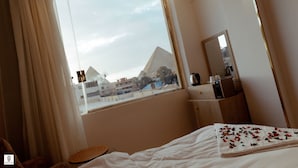 Une chambre d’hôtel avec une grande fenêtre, un lit, une table de chevet en bois, un miroir et une vue sur une pyramide.