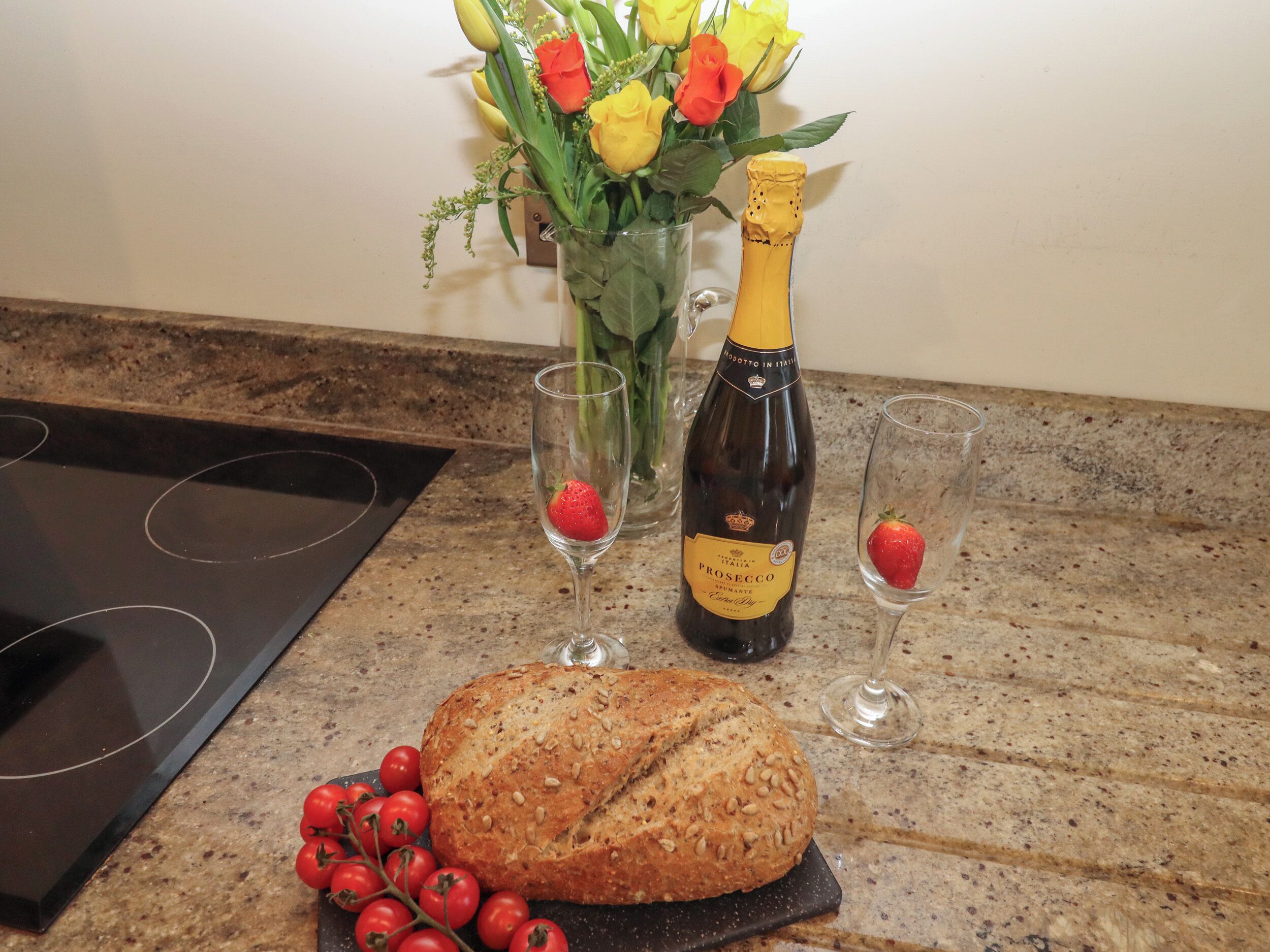A kitchen counter with a loaf of bread, two glasses with strawberries, a bottle of Prosecco, and a vase with flowers.
