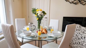A dining area with a glass table surrounded by white chairs, a vase of flowers, breakfast items, and a fireplace in the background. - Powder Keg (Whitby)