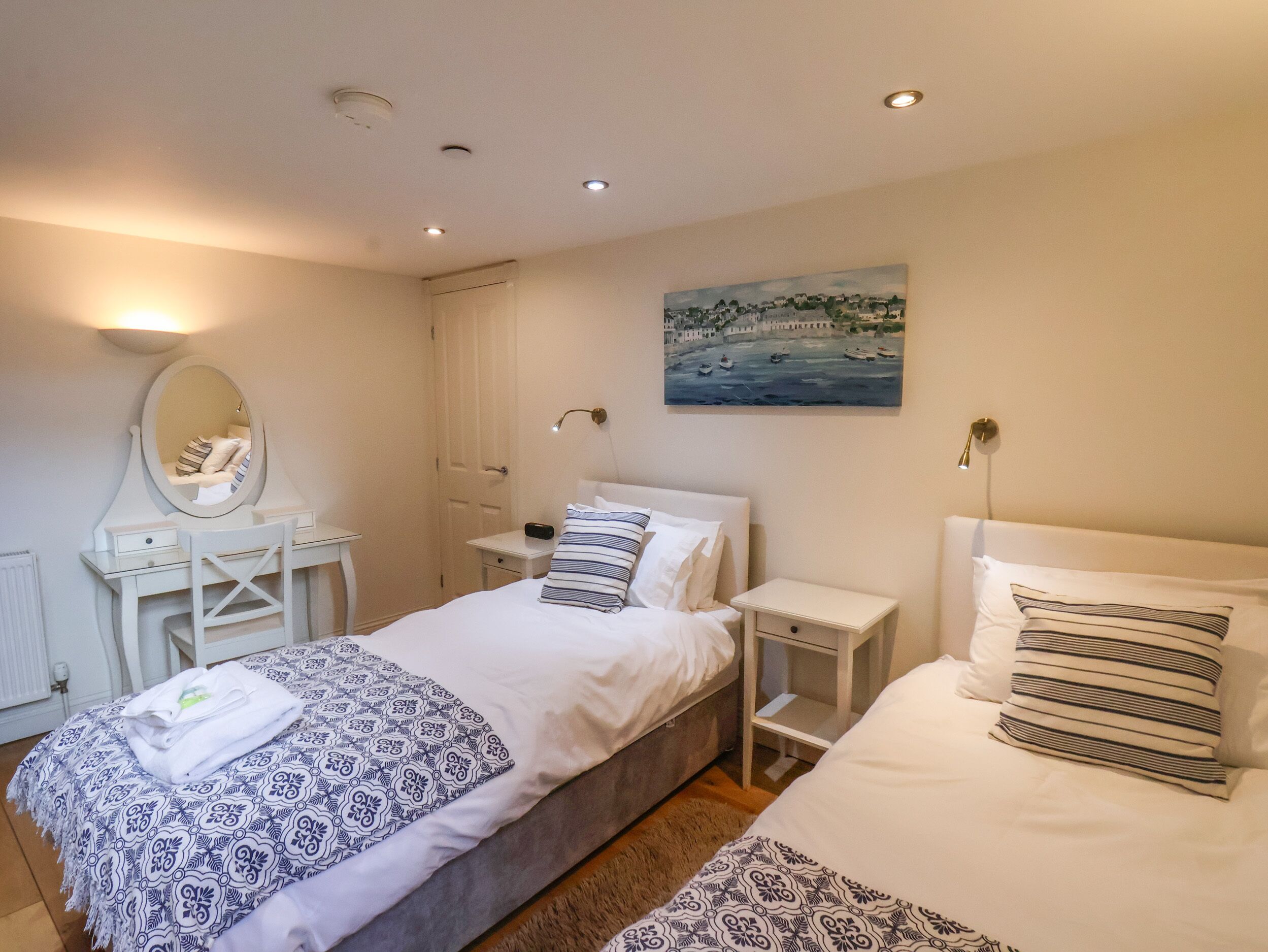 Un dormitorio con dos camas, una mesita de noche con lámpara, un tocador con espejo y un cuadro en la pared.