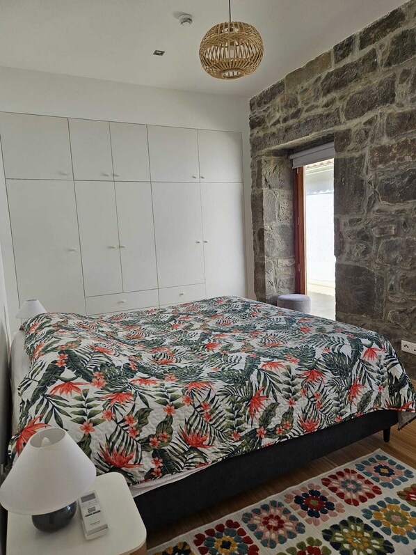 Une chambre à coucher avec un lit, une armoire blanche, un mur en pierre, un luminaire suspendu et une fenêtre donnant sur un paysage.