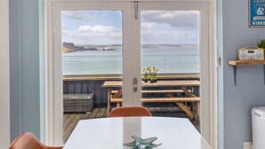 A room with a large window overlooking a deck with wooden furniture and a view of the sea.