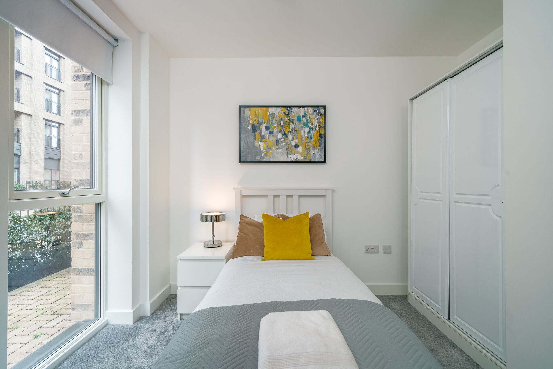 Una camera da letto con un letto, un comodino, una lampada, un quadro e vista sull'esterno.