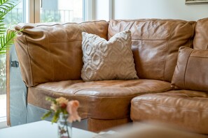 A brown leather sofa with a single white cushion, positioned near a window with a view of greenery outside.