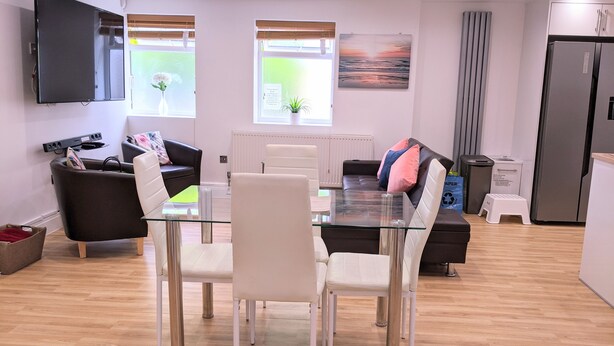 Een moderne woonkamer met een glazen eettafel, witte stoelen, een zwarte bank en een grote flatscreen televisie aan de muur.