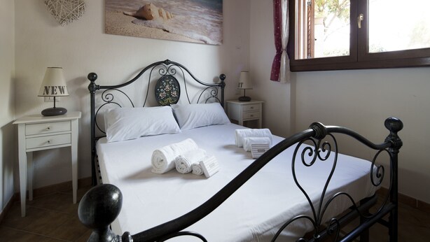 A neatly made bed with white linens, two bedside tables with lamps, and a painting of a beach scene on the wall. - Casa Armonia – Terrace & Beach Nearby | Welcomely (Tanaunella)
