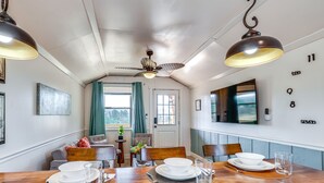 Un coin repas avec une table en bois dressée pour quatre personnes, un ventilateur de plafond et un téléviseur fixé au mur.