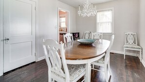 A dining room with a wooden table, white chairs, a chandelier, and a window with blinds.