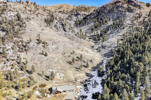 Un chalet doté d’un toit en métal et de murs en pierre, entouré d’un paysage montagneux parsemé de quelques arbres et de plaques de neige.