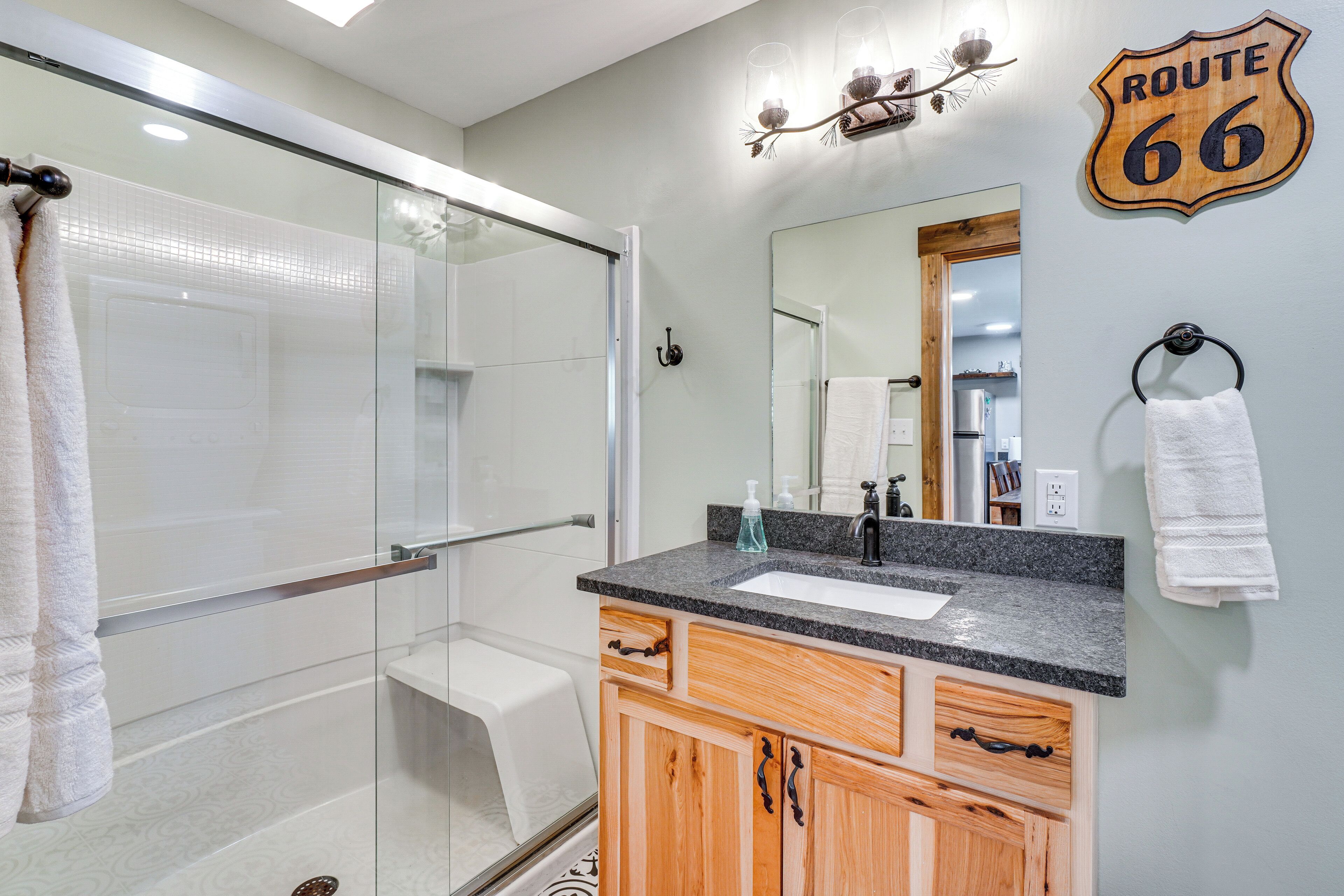 Un baño con cabina de ducha de vidrio, lavamanos con encimera de granito, armarios de madera y un espejo con un cartel de 'Route 66'.