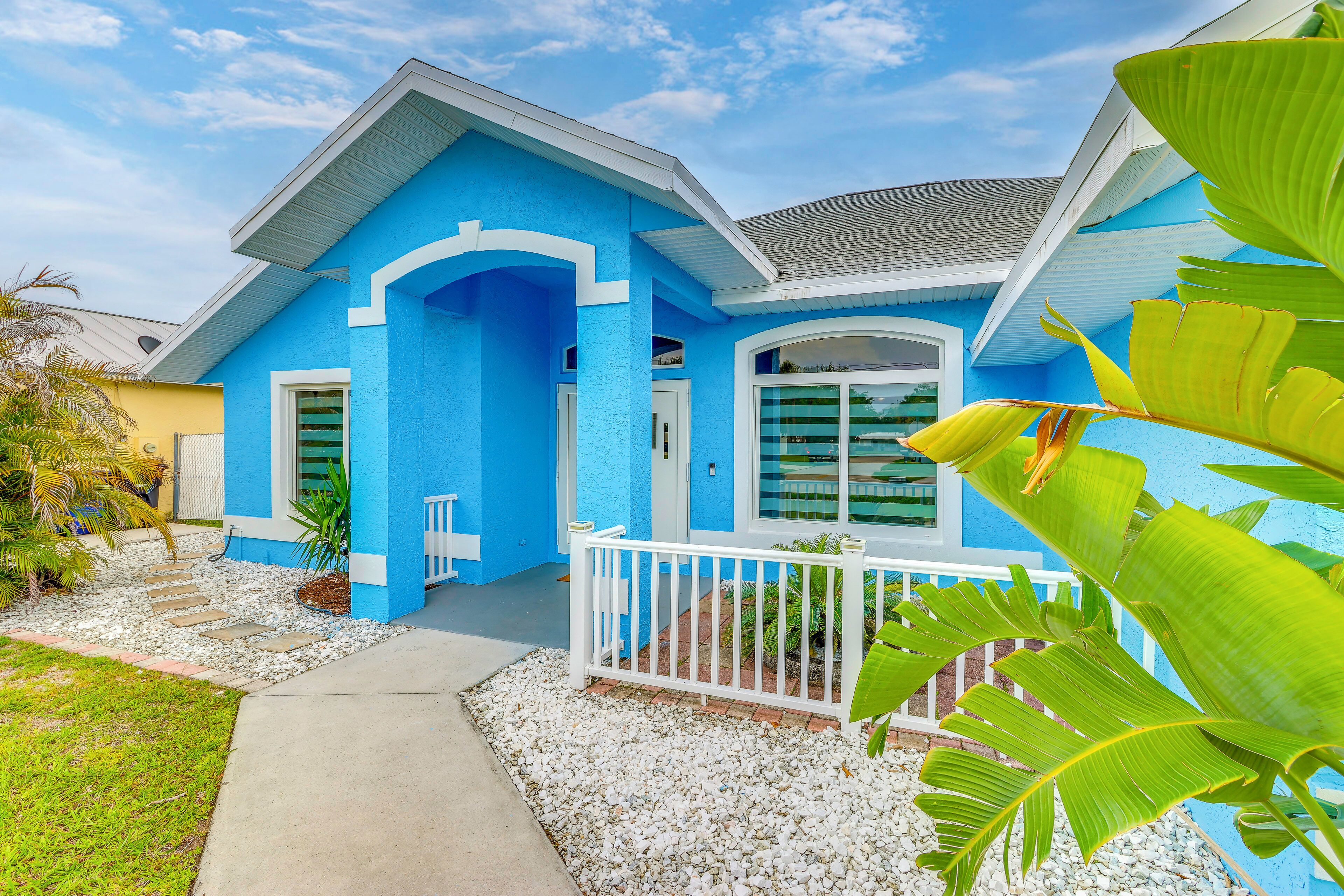 Una casa blu con un portico bianco, un vialetto e piante tropicali.