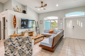 A living room with a sofa, coffee table, and TV. There is a ceiling fan, a large window, and a door.