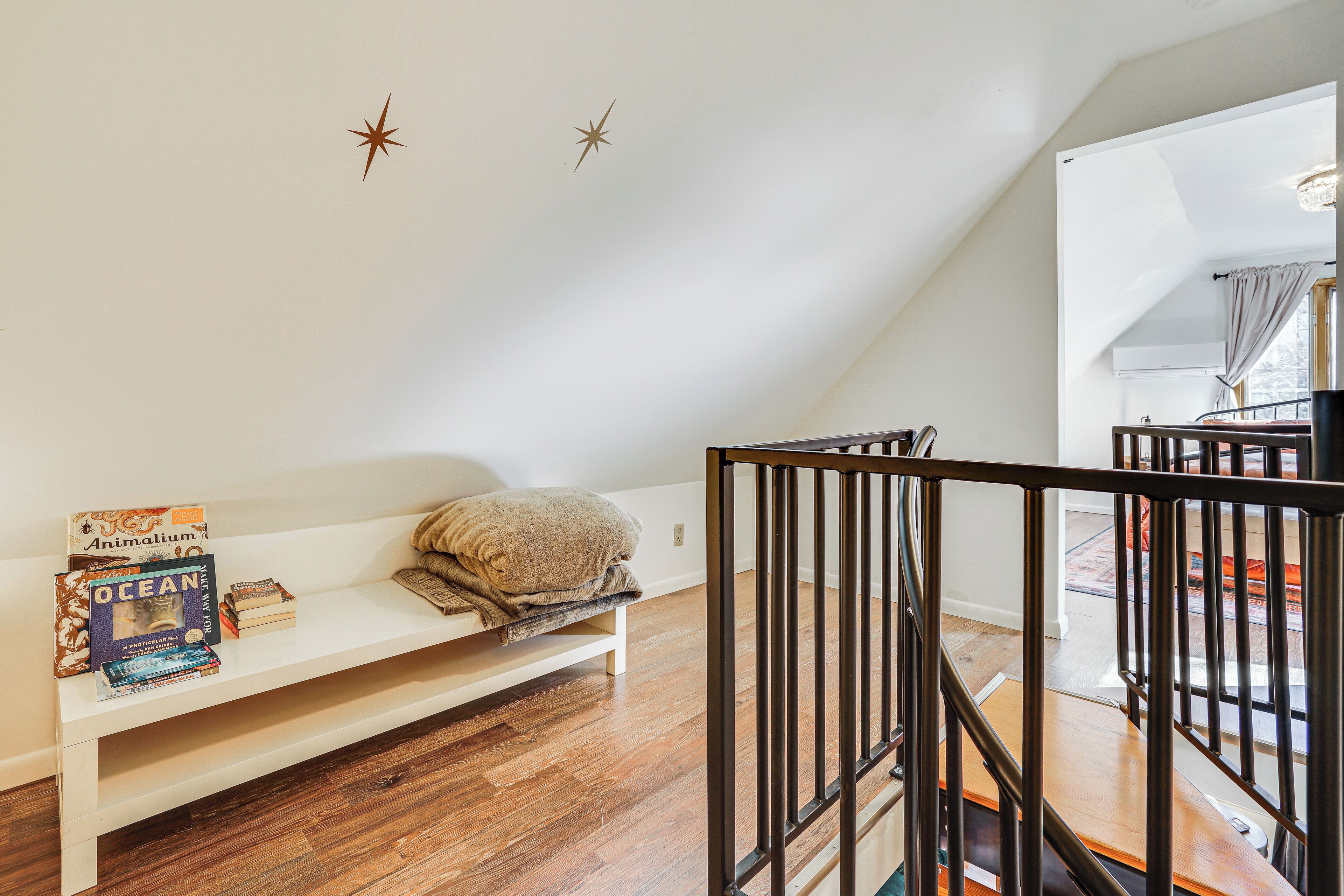 A room with a wooden floor, a white bench with a cushion, and a staircase with a black railing.