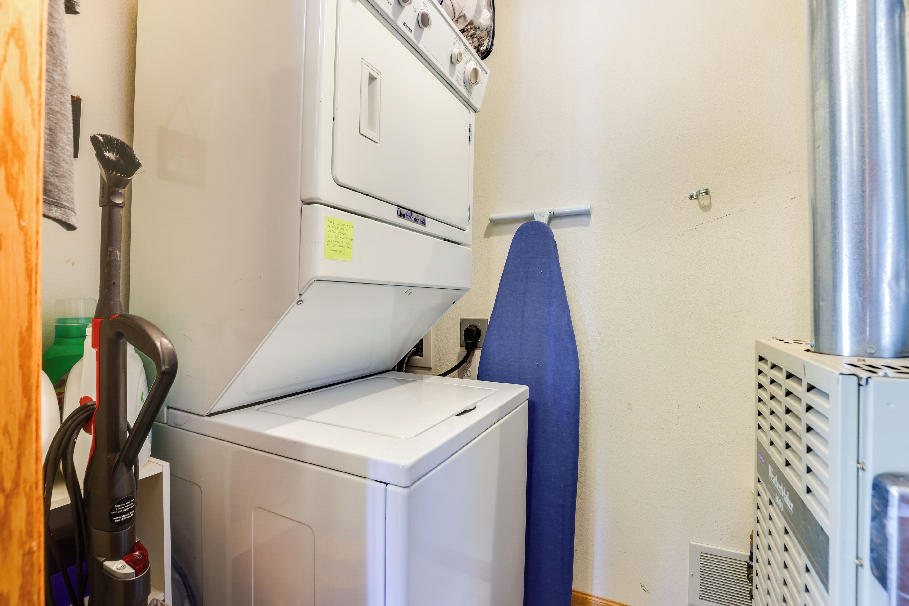 A laundry area with a front-loading washer and dryer, a blue ironing board, and a vacuum cleaner.