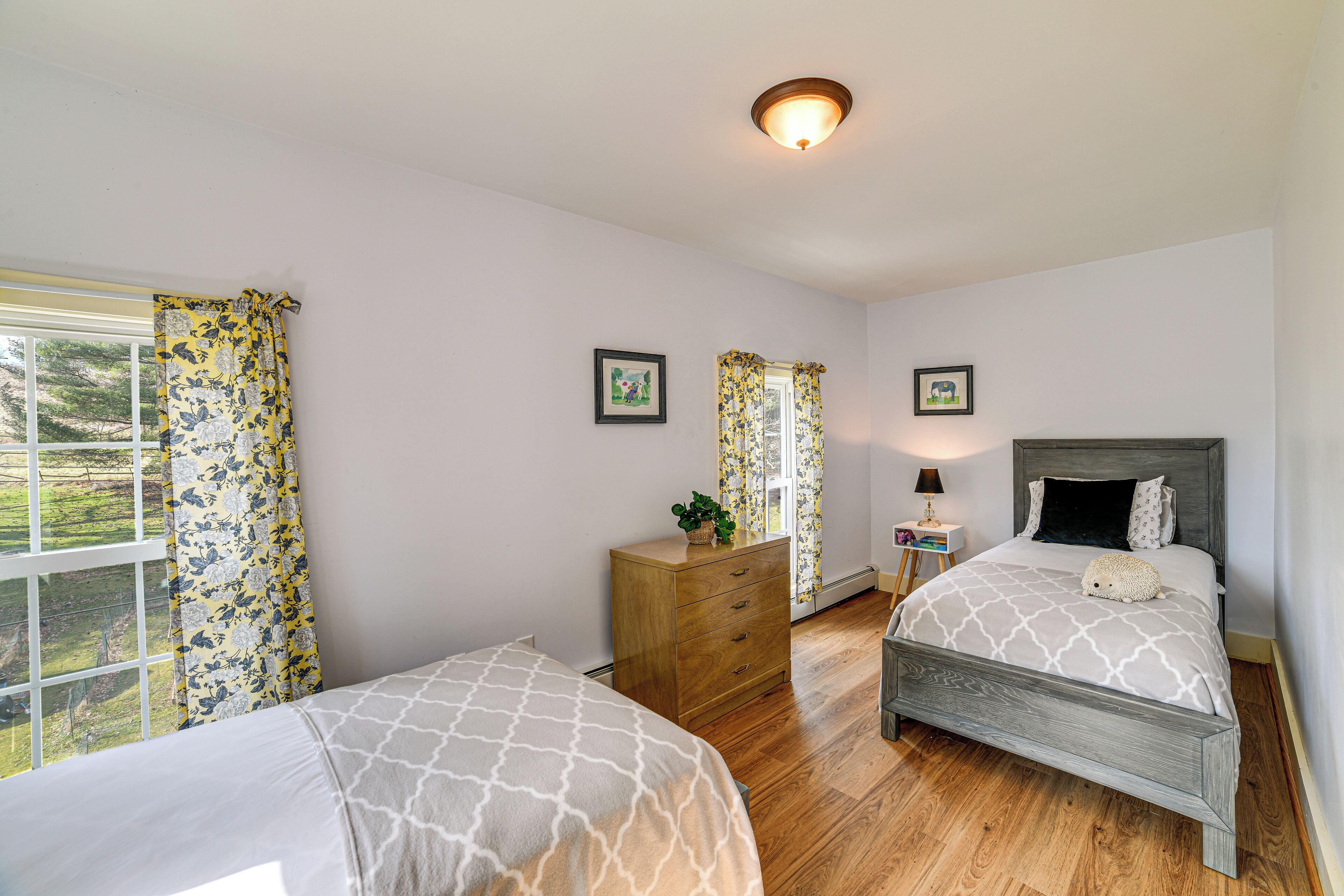 A bedroom with two beds, wooden flooring, a dresser, and a window with floral curtains.