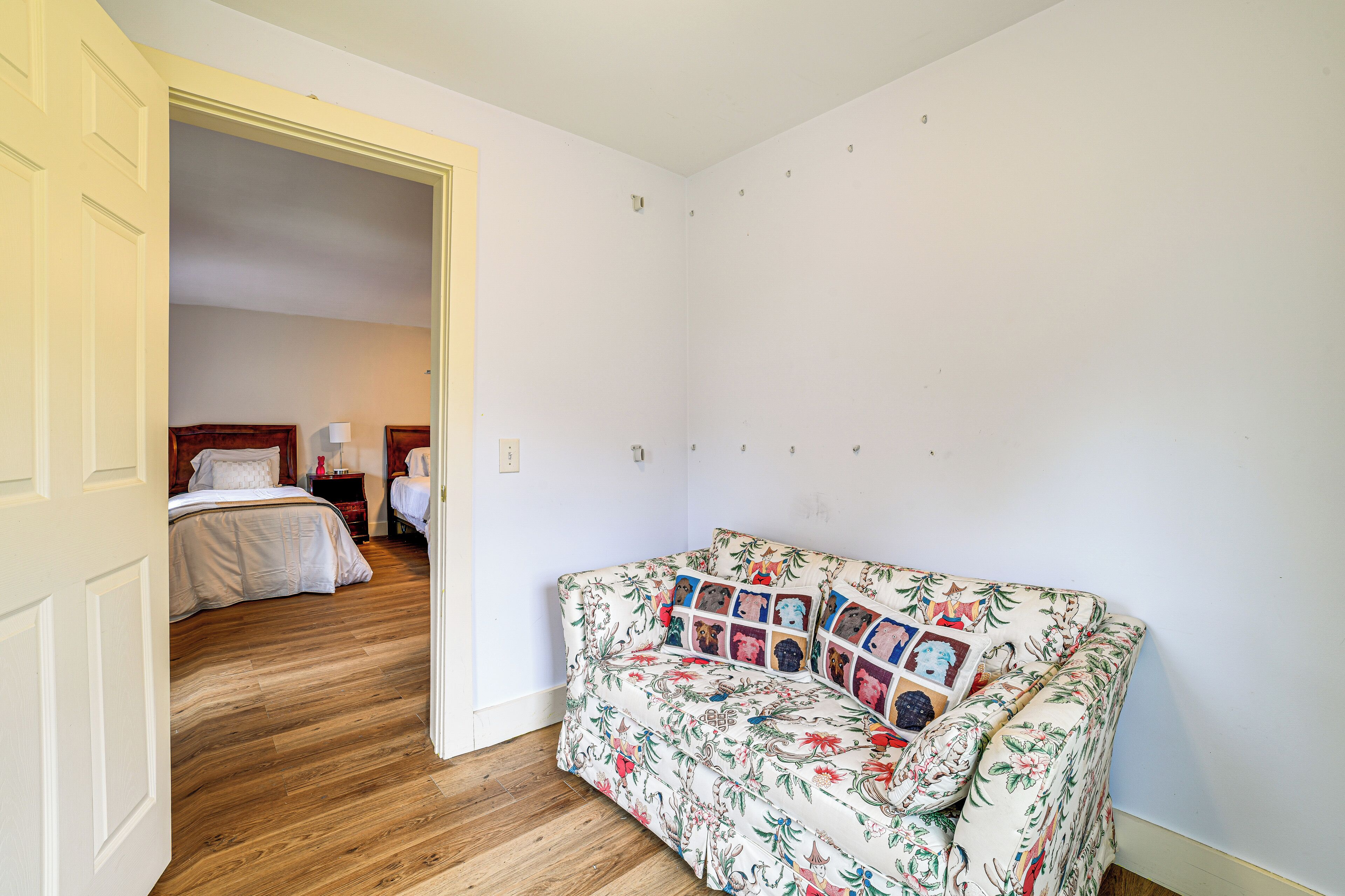 A room with a wooden floor, a floral sofa, a bed with white bedding, and a nightstand with a lamp.