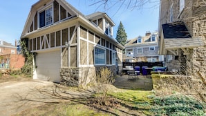A two-story house with a garage, stone walls, and a yard with a fence and outdoor furniture. - Cozy Niagara Falls Cottage w/ Gas Grill + Patio! (Niagara Falls)