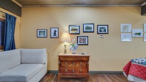 Una habitación con pared beige, suelo de madera, un sofá blanco, un aparador de madera con cajones, una lámpara y cuadros en la pared.