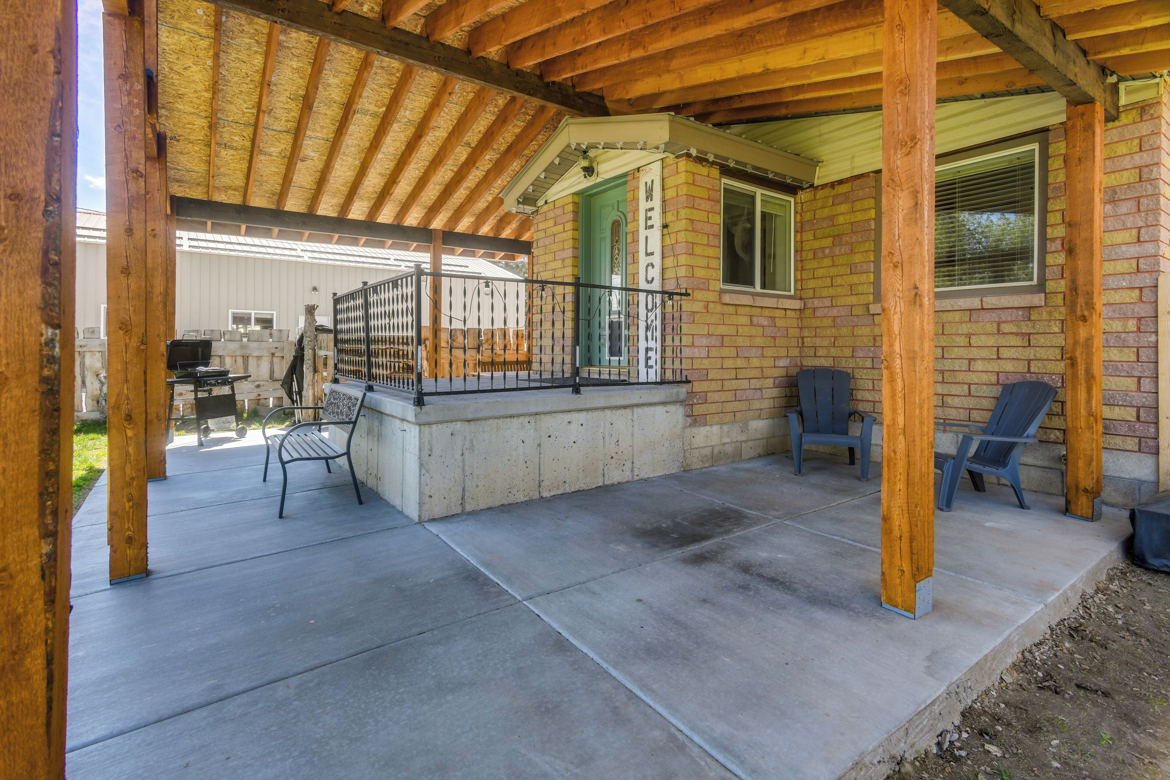 A covered patio area with a concrete floor, wooden beams, and a brick wall. There are two chairs and a bench.