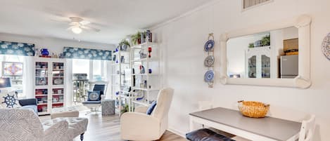 A spacious living room with a white sofa, a white armchair, a dining table with a basket, and a bookshelf filled with decorative items.