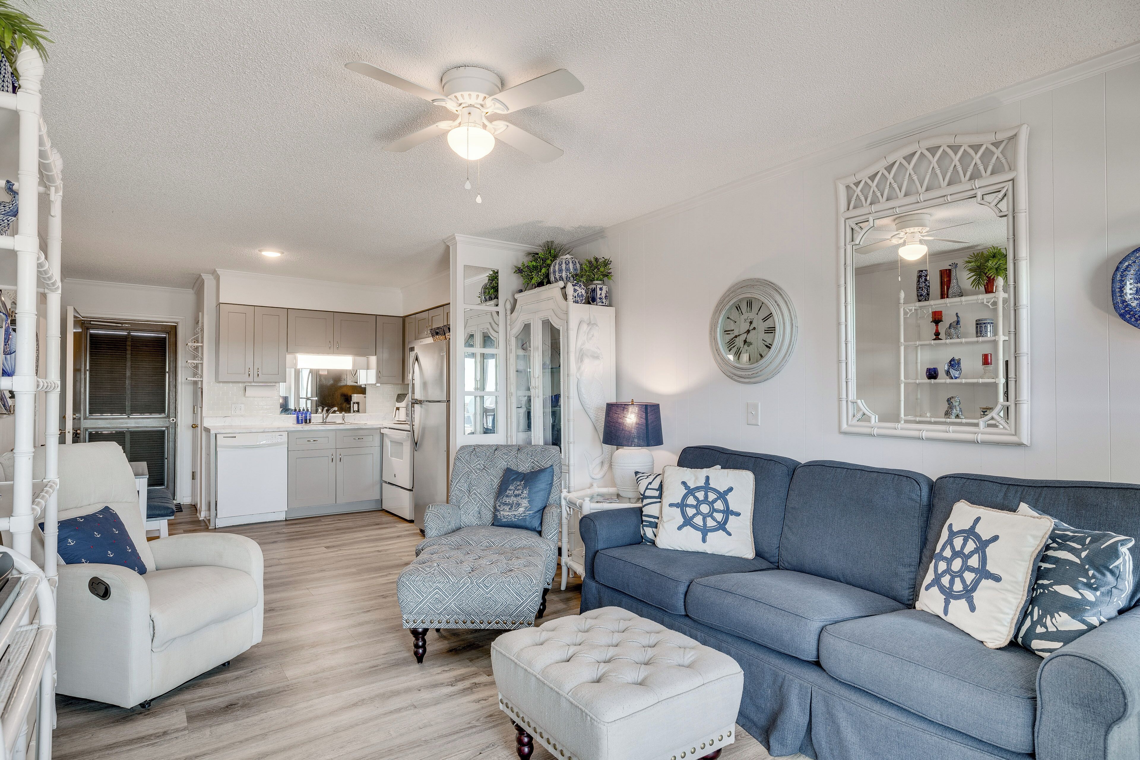A modern living room with a blue sofa, a white armchair, and a matching ottoman. There is a dining area in the background with a kitchen visible.