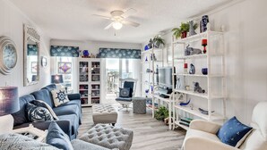 A living room with a white ceiling fan, a blue sofa, a white armchair, and a bookshelf with decorative items. - Atlantic Beach Condo w/ Balcony & Beach Access! (Atlantic Beach)