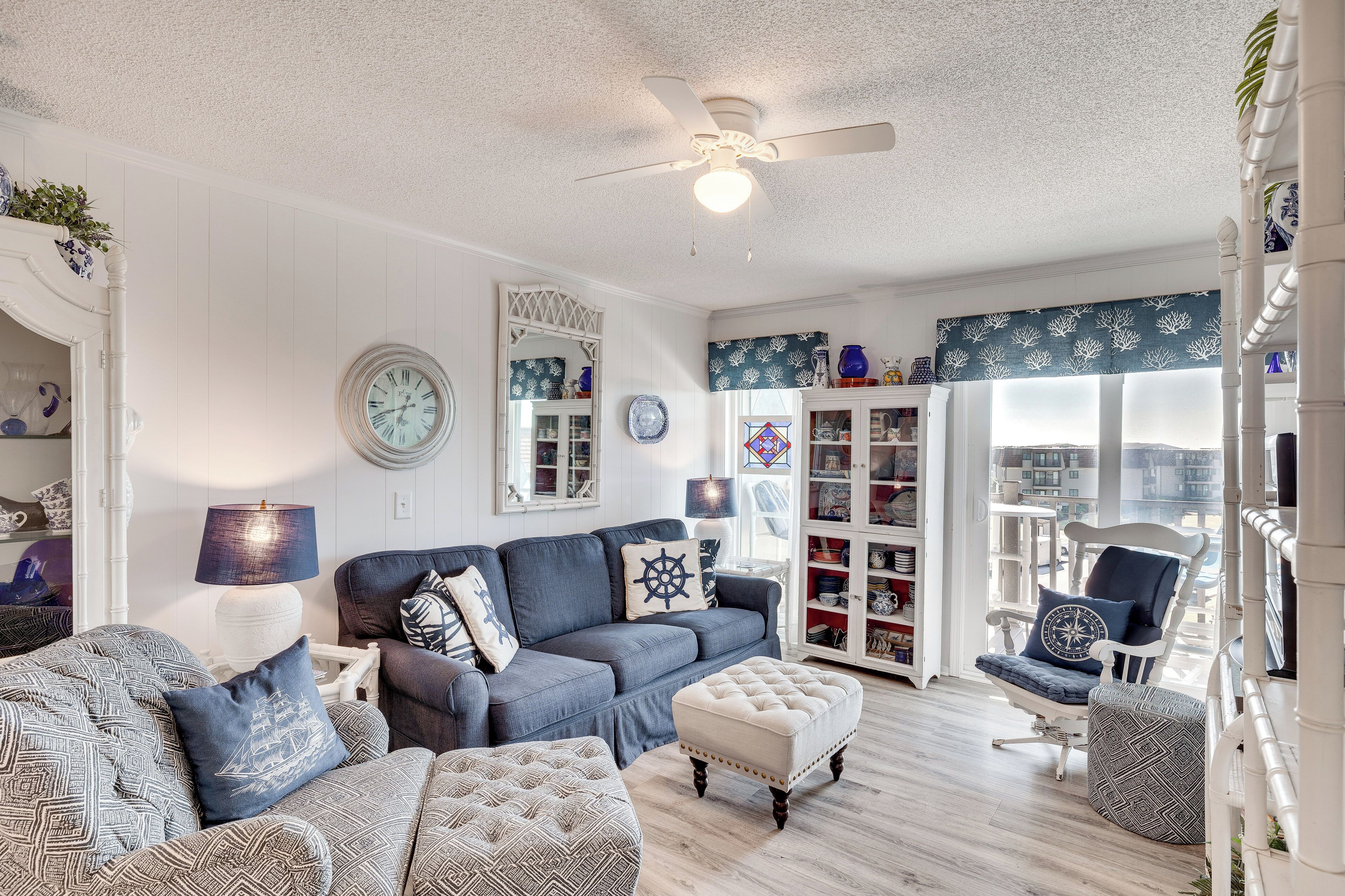 A living room with a blue sofa, a white armchair, and a matching ottoman. There is a ceiling fan, a bookshelf, and a window with a view.