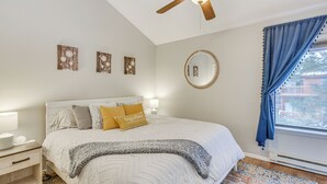 Quarto com cama, mesas de cabeceira, ventoinha no teto, janela com cortinas e decoração nas paredes.