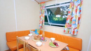 A dining area with a table set for two, a wicker basket, a bottle of juice, and a bowl of fruit. - Mobil-home Classique 2 Chambres (Telgruc-sur-Mer)