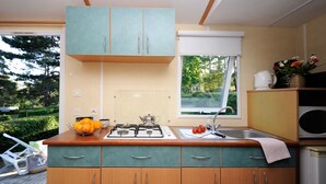 A compact kitchen with teal cabinets, a gas stove, a sink, and a window offering a view of greenery. - Mobil-home Classique 2 Chambres (Telgruc-sur-Mer)
