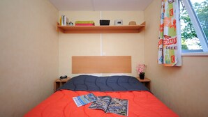 A bedroom with a single bed, a wooden headboard, a shelf above the bed, and a window with curtains. - Mobil-home Classique 2 Chambres (Telgruc-sur-Mer)