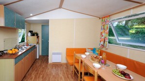 A compact kitchen with a dining area, wooden cabinets, and a window with curtains. - Mobil-home Classique 2 Chambres (Telgruc-sur-Mer)