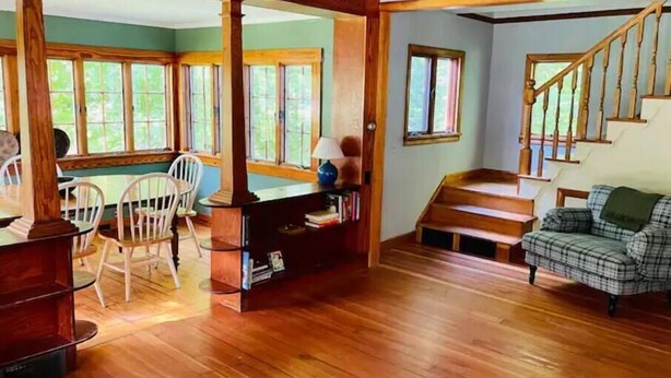 A spacious living room with a wooden staircase, a dining area with chairs, and a cozy seating area with a plaid sofa. - Sweet comfy cottage on Small Point Road (Phippsburg)