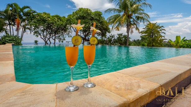 Vista a la piscina con dos vasos de bebida de naranja, palmeras y cielo despejado.