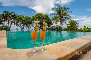 Une vue sur la piscine avec deux verres d’une boisson orange, des palmiers et un ciel dégagé.